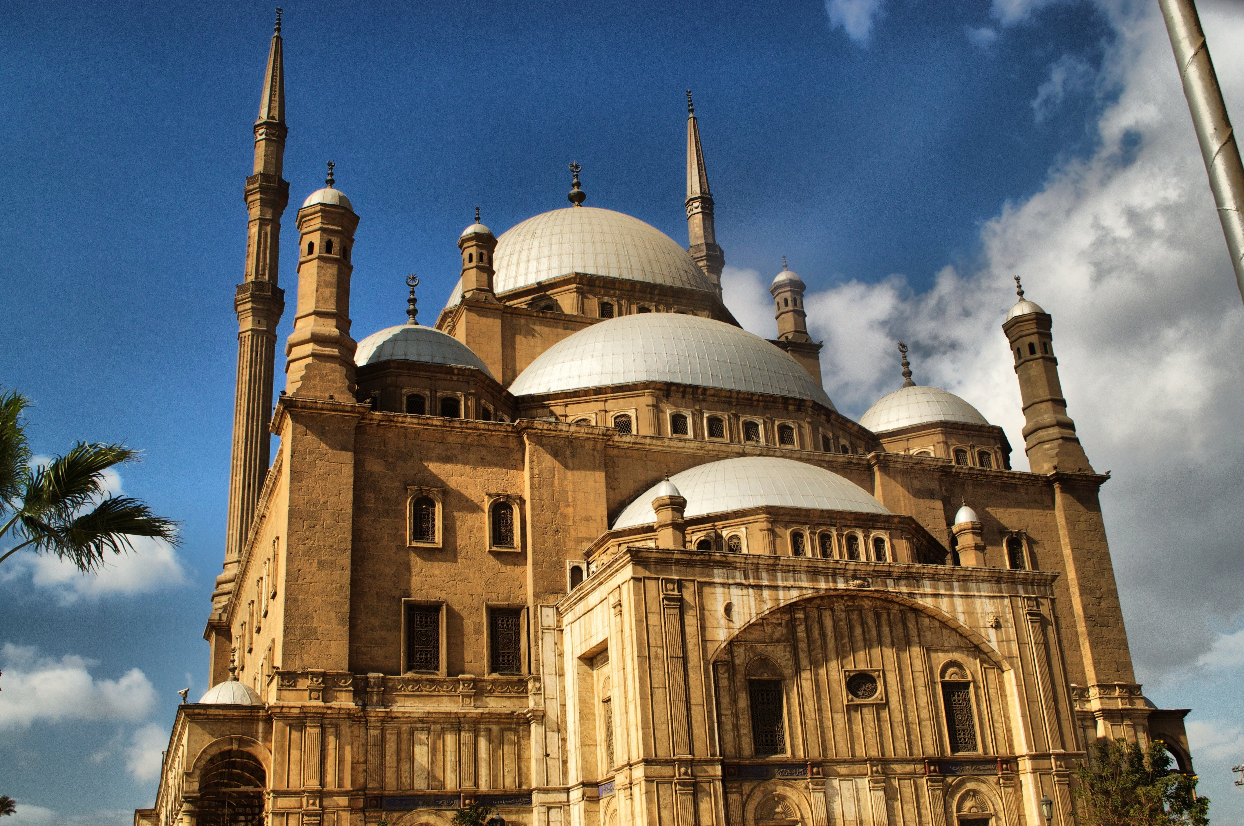 Mohamed Ali Mosque is amongst the most interesting Mosques in Egypt. It stands proudly on the highest point inside the courtyard of the Citadel of Saladin, and is also called the Alabaster Mosque. The architect was Yousf Boushnaq, a Turkish man who had come over from Istanbul to build this great Mosque for Mohamed Ali, the ruler of Egypt from1805 until 1849. He based his plans on the Sultan Ahmed Mosque in Istanbul, and the construction began in 1830 A.D. The work continued ceaselessly until the death of Mohamed Ali in 1849, and had to be finished during the reign of his successors. Mohamed Ali was buried in the tomb situated on the south-eastern side of Beit Al Salah, on the right side of the entrance that leads to the main section. In 1899 the Mosque showed signs of cracking and repairs were undertaken, but some of these repairs were not adequate. Therefore, in 1931, during the reign of King Fuad I, a committee was set up, comprising of several great architects, which eventually presented a report recommending the demolition of the big main dome, the semi domes and the small domes, and then reconstructing them according to the original design. Between 1931 and 1939, the project, including demolition, building and rebuilding, painting and gilding, was undertaken; the total cost being 100,000 LE. The main material used for the construction was limestone, but the lower parts of the Mosque, and the forecourt, are faced to a height of 11.5m with alabaster. The Mosque is rectangular in shape and consists of two sections: The Eastern Section, which is the main section, called “Beit al Salah” or “House of Prayer”. The Western Section, called the “Sahn” “or “Courtyard”. The Eastern Section (The “Beit al Salah” or “House of Prayer”) The eastern section is the part that was dedicated to prayer. It is square in shape, each side measure 41m, and has a roof with a central dome (52m in height) resting on four large arches supported by massive piers. Surrounding the big central dome there are four half domes, while there are four more small domes covering the corners. The marble mihrab is covered by a half-dome at the lower level. The domes are pointed and covered with medallions and other motifs. The interior dome is impressive because of its size and shape, similar to the Mosques of Istanbul. There are 6 medallions around the dome, which include the names of Allah (God) and Mohamed (the Prophet), as well as the names of the four rightly guided Caliphs, namely Abou Bakr, Omar, Othman, and Ali. The mosque has 2 Minbars or pulpits; the original one is the larger, it is made of wood decorated with gilded ornaments, while the smaller one is of marble, it was gifted to the mosque by king Farouk in 1939 A.D. Above the entrance is a grand gallery supported on marble pillars with bronze balustrade. To the right of the entrance is the tomb of Mohamed Ali. It is made white marble covered with floral motifs, and pointed and gilded inscriptions. originally Mohamed Ali was not buried in his mosque but later during the time king Abbas I (1849-1854), His body was transferred from Housh El Basha to the inside of the mosque where it rests inside The bronze grill. The Western Section (The Courtyard or the Sahn) It is a large open courtyard of about 54 m in length and 53 m in width. It is surrounded by a single arched riwaqs or naves raised on pillars and roofed with small domes. In the middle of the courtyard there is the ablution fountain, it is octagonal in shape and covered by a large leaded domed canopy resting on 8 pillars with natural ornaments. Inside the dome is another marble small dome and it is octagonal in shape, decorated with floral motifs. In the walls of the riwaqs of the courtyard there are 46 widows. While the Eastern wall which overlooks the Eastern Section, it has 8 windows above which there is a frieze of inscription of the Koran (Surat Al Fath), Above the entrance to the Easter section there is frieze that bears the name of the Turkish sultan Abd Al Maguid. Opposite to the doorway of the prayer House, at the far end of the centre of the NW Riwaq is a pavilion, above which is an elaborate French Clock, presented to Mohamed Ali in 1845 by the King Luis Philip in exchange of the obelisk which is now standing in the Concorde square in Paris. This clock has never been working properly! At the west and the North Corners are 2 slender octagonal minarets that rise to 82 M in height. And has with 2 balconies.