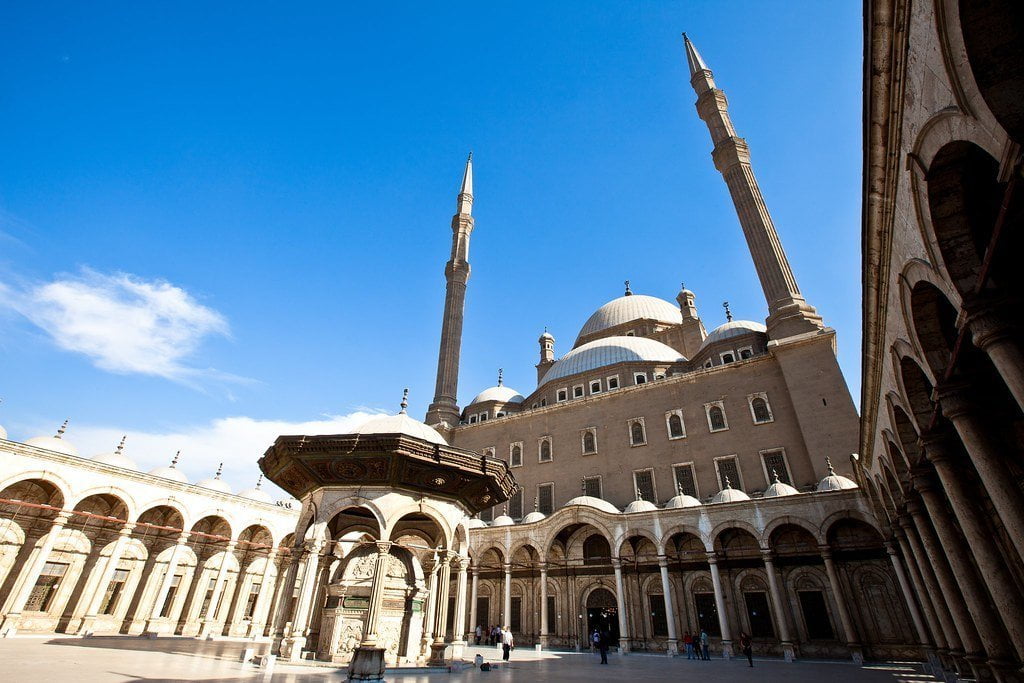 Mohamed Ali Mosque is amongst the most interesting Mosques in Egypt. It stands proudly on the highest point inside the courtyard of the Citadel of Saladin, and is also called the Alabaster Mosque. The architect was Yousf Boushnaq, a Turkish man who had come over from Istanbul to build this great Mosque for Mohamed Ali, the ruler of Egypt from1805 until 1849. He based his plans on the Sultan Ahmed Mosque in Istanbul, and the construction began in 1830 A.D. The work continued ceaselessly until the death of Mohamed Ali in 1849, and had to be finished during the reign of his successors. Mohamed Ali was buried in the tomb situated on the south-eastern side of Beit Al Salah, on the right side of the entrance that leads to the main section. In 1899 the Mosque showed signs of cracking and repairs were undertaken, but some of these repairs were not adequate. Therefore, in 1931, during the reign of King Fuad I, a committee was set up, comprising of several great architects, which eventually presented a report recommending the demolition of the big main dome, the semi domes and the small domes, and then reconstructing them according to the original design. Between 1931 and 1939, the project, including demolition, building and rebuilding, painting and gilding, was undertaken; the total cost being 100,000 LE. The main material used for the construction was limestone, but the lower parts of the Mosque, and the forecourt, are faced to a height of 11.5m with alabaster. The Mosque is rectangular in shape and consists of two sections: The Eastern Section, which is the main section, called “Beit al Salah” or “House of Prayer”. The Western Section, called the “Sahn” “or “Courtyard”. The Eastern Section (The “Beit al Salah” or “House of Prayer”) The eastern section is the part that was dedicated to prayer. It is square in shape, each side measure 41m, and has a roof with a central dome (52m in height) resting on four large arches supported by massive piers. Surrounding the big central dome there are four half domes, while there are four more small domes covering the corners. The marble mihrab is covered by a half-dome at the lower level. The domes are pointed and covered with medallions and other motifs. The interior dome is impressive because of its size and shape, similar to the Mosques of Istanbul. There are 6 medallions around the dome, which include the names of Allah (God) and Mohamed (the Prophet), as well as the names of the four rightly guided Caliphs, namely Abou Bakr, Omar, Othman, and Ali. The mosque has 2 Minbars or pulpits; the original one is the larger, it is made of wood decorated with gilded ornaments, while the smaller one is of marble, it was gifted to the mosque by king Farouk in 1939 A.D. Above the entrance is a grand gallery supported on marble pillars with bronze balustrade. To the right of the entrance is the tomb of Mohamed Ali. It is made white marble covered with floral motifs, and pointed and gilded inscriptions. originally Mohamed Ali was not buried in his mosque but later during the time king Abbas I (1849-1854), His body was transferred from Housh El Basha to the inside of the mosque where it rests inside The bronze grill. The Western Section (The Courtyard or the Sahn) It is a large open courtyard of about 54 m in length and 53 m in width. It is surrounded by a single arched riwaqs or naves raised on pillars and roofed with small domes. In the middle of the courtyard there is the ablution fountain, it is octagonal in shape and covered by a large leaded domed canopy resting on 8 pillars with natural ornaments. Inside the dome is another marble small dome and it is octagonal in shape, decorated with floral motifs. In the walls of the riwaqs of the courtyard there are 46 widows. While the Eastern wall which overlooks the Eastern Section, it has 8 windows above which there is a frieze of inscription of the Koran (Surat Al Fath), Above the entrance to the Easter section there is frieze that bears the name of the Turkish sultan Abd Al Maguid. Opposite to the doorway of the prayer House, at the far end of the centre of the NW Riwaq is a pavilion, above which is an elaborate French Clock, presented to Mohamed Ali in 1845 by the King Luis Philip in exchange of the obelisk which is now standing in the Concorde square in Paris. This clock has never been working properly! At the west and the North Corners are 2 slender octagonal minarets that rise to 82 M in height. And has with 2 balconies.